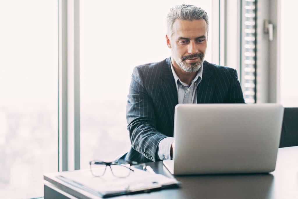 A businessman typing on a laptop in an office while sitting at a desk with glass and a notebook beside him.
