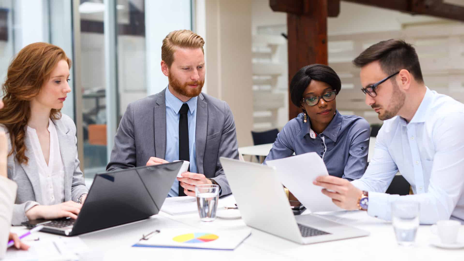 A marketing team sitting around a table doing a competitive research meeting with laptops and papers.